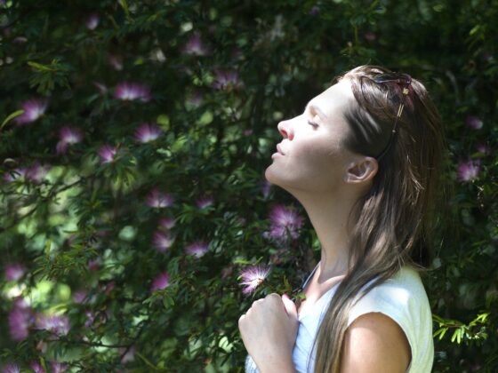woman closing her eyes against sun light standing near purple petaled flower plant